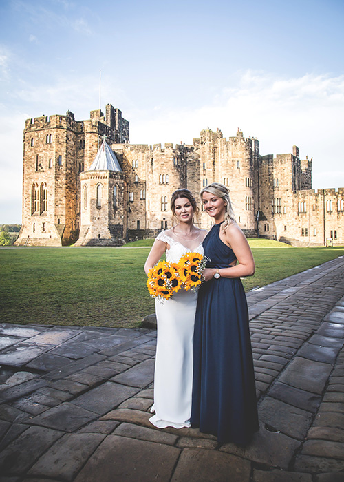 bride and bridesmaid in front of alnwick castle blue sky